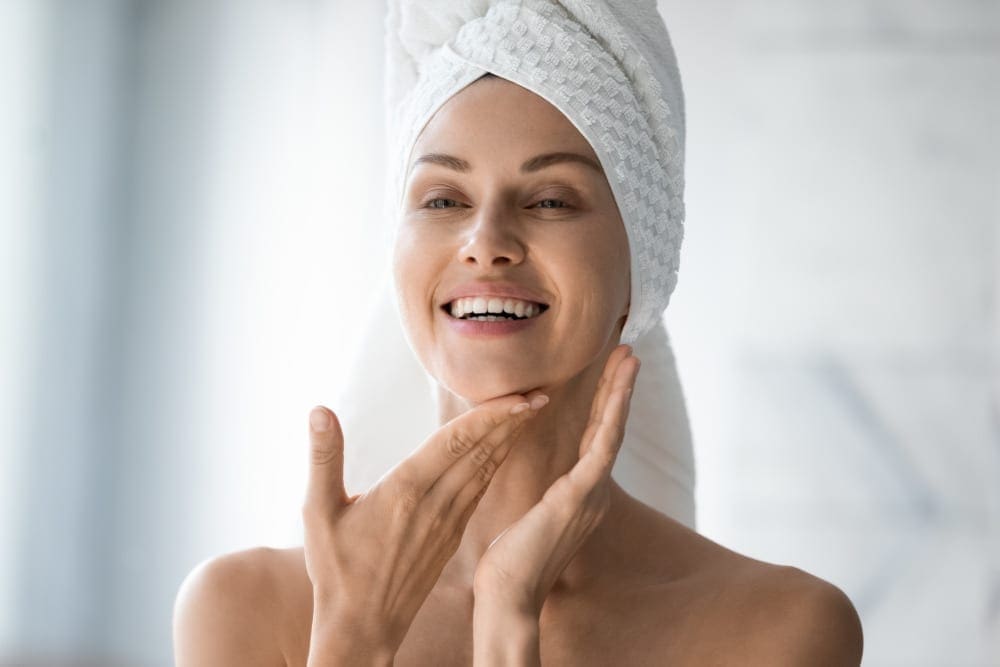 Happy woman with her hair wrapped in a towel looking at her liquid facelift results in the bathroom mirror.